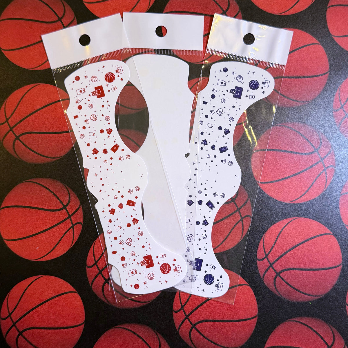 Three white novelty sock-shaped stickers or decals in clear plastic sleeves, decorated with red and blue basketball-themed patterns, displayed on a background of red basketballs.